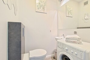A modern bathroom with a grey cabinet, a white toilet, a washing machine, and framed botanical artwork on the walls.