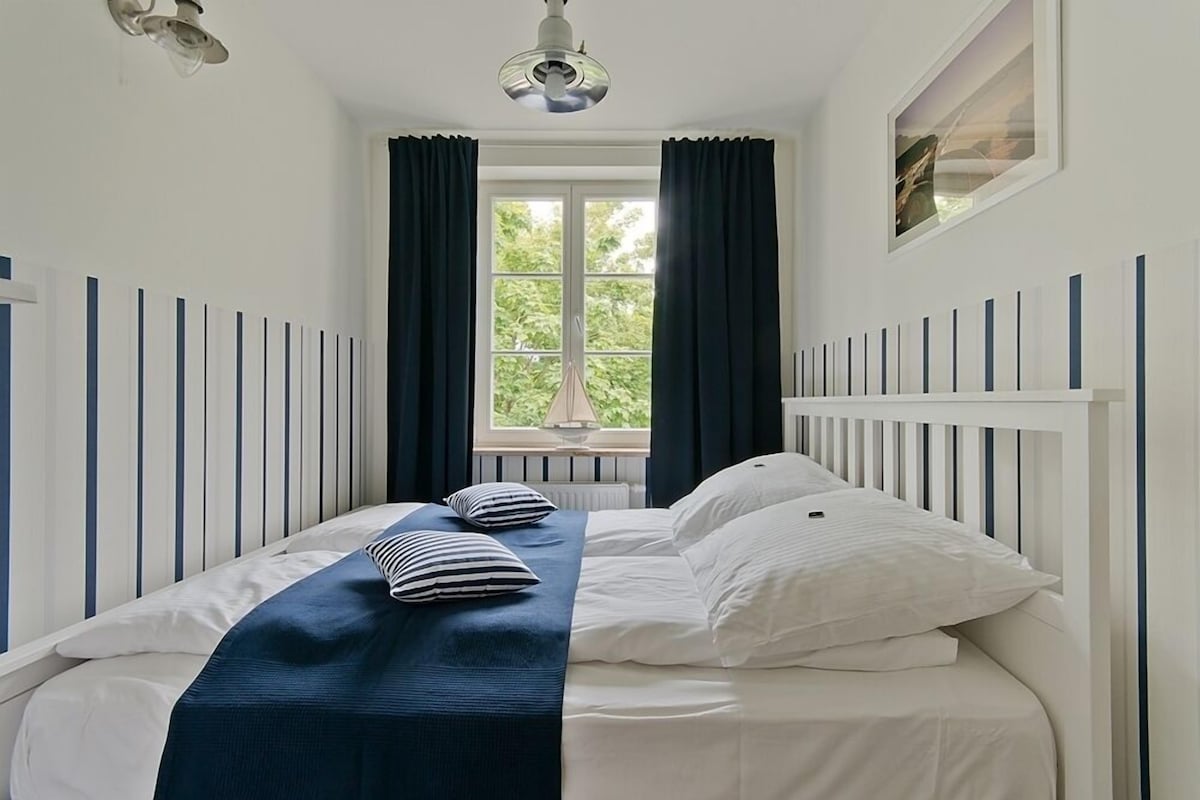 A neatly made bed with a blue bedspread and striped pillows, a window with curtains, and a ceiling fan.
