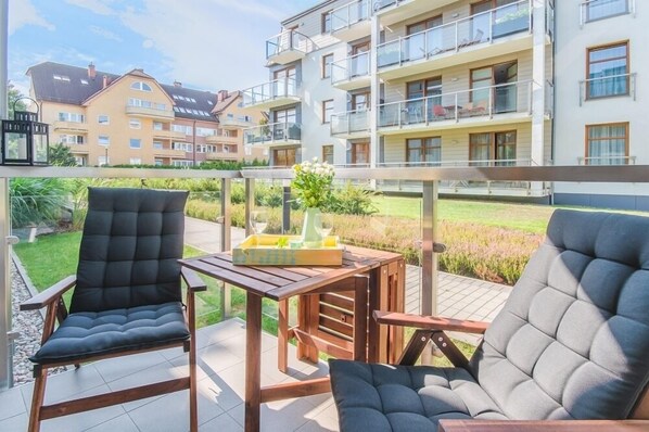 A balcony with two black cushioned chairs and a wooden table, overlooking a residential area with multi-story buildings.