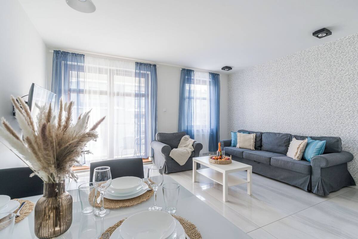 Un salon moderne avec un canapé gris, une table basse blanche et un coin repas avec une table en verre, mise pour quatre personnes.