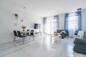 A modern living room with a dining area, large windows with blue curtains, and a flat-screen TV mounted on the wall. - Imperial Apartments - Sopocka Przystan (Sopot)