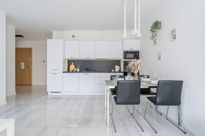 A modern kitchen with white cabinets, a black countertop, and stainless steel appliances. There is a dining area with a glass table and black chairs. - Imperial Apartments - Sopocka Przystan (Sopot)