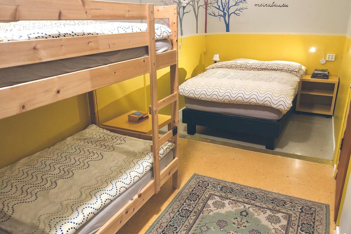 A bunk bed with a patterned bedspread, a bedside table with a lamp, and a wall with a tree mural.