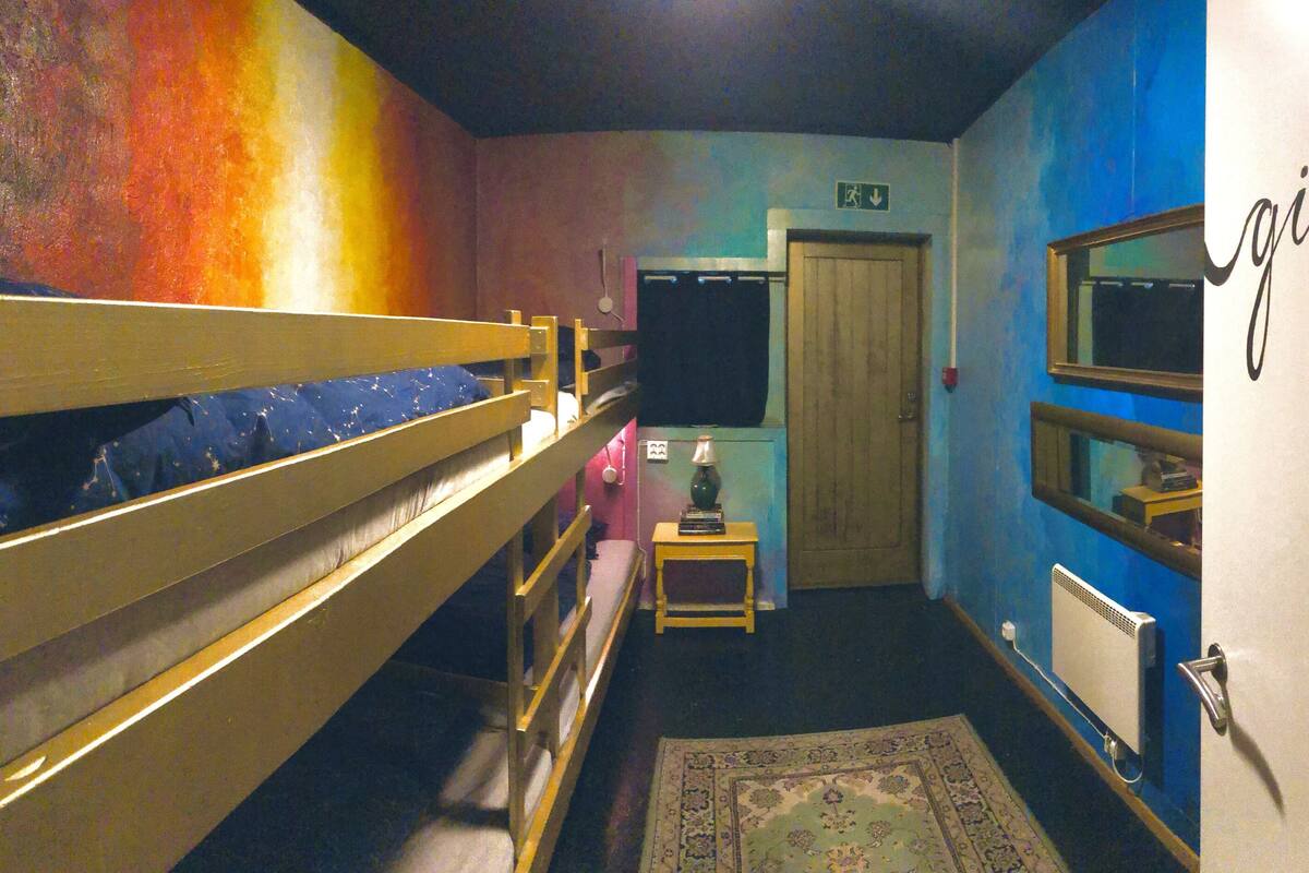 A dormitory room with bunk beds, a small table with a lamp, a radiator, and a door with a sign.