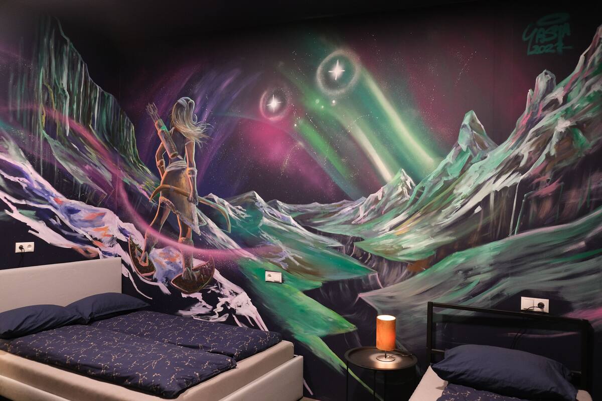 A room with a bed, a small table with a lamp, and a mural of a cosmic scene with a figure.