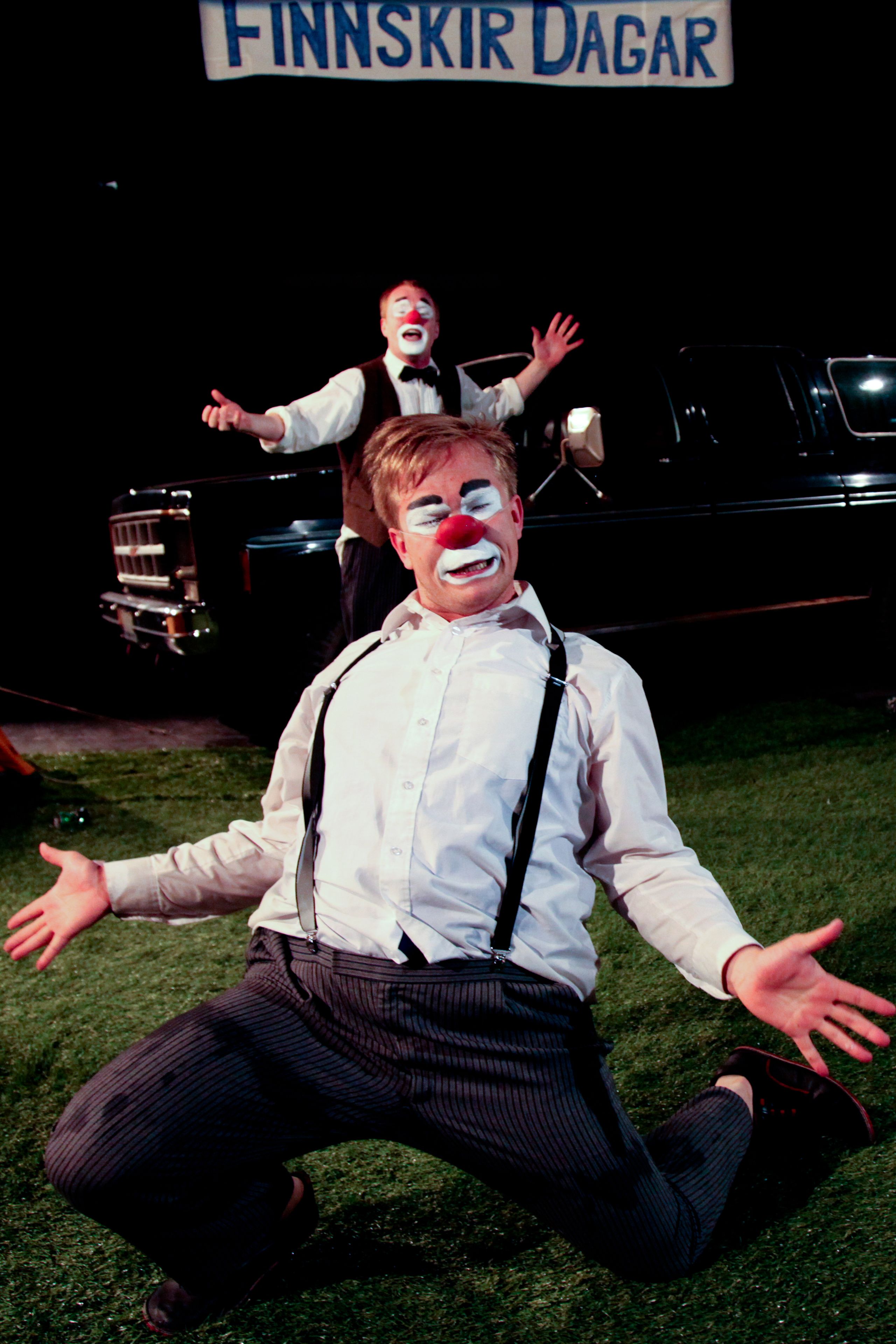 A person in a white shirt and black suspenders kneeling on grass with arms outstretched.