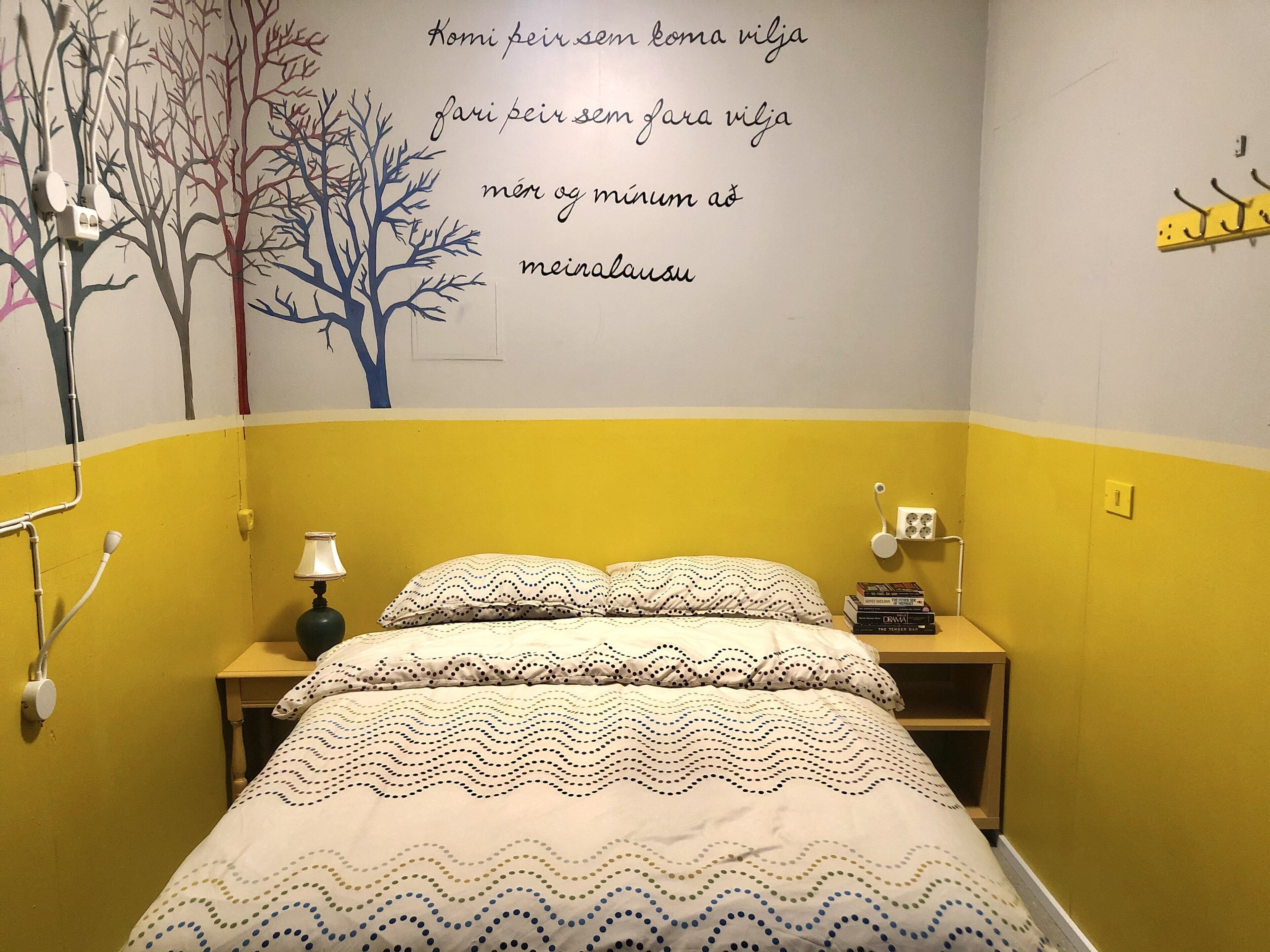 Een slaapkamer met een bed, nachtkastje, lamp en een muur met een boommuurcitatie en tekst.