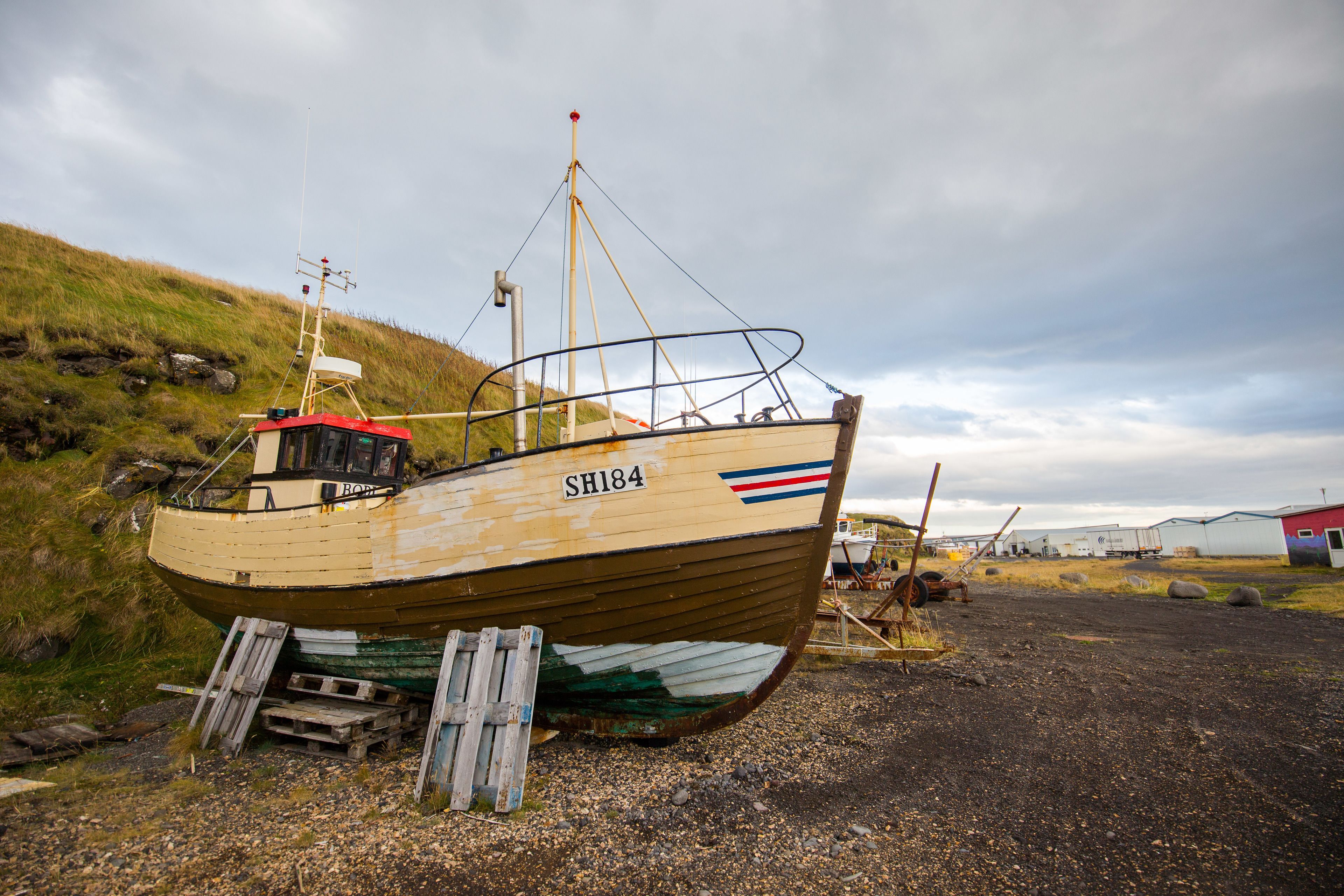 A stationary boat with the identifier SH184, docked on a gravel surface near a grassy hill and a building.