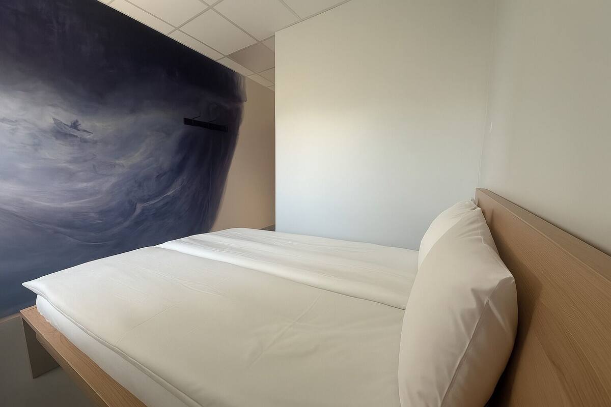A bed with a wooden headboard and a white mattress in a room with a plain wall and a large abstract painting.