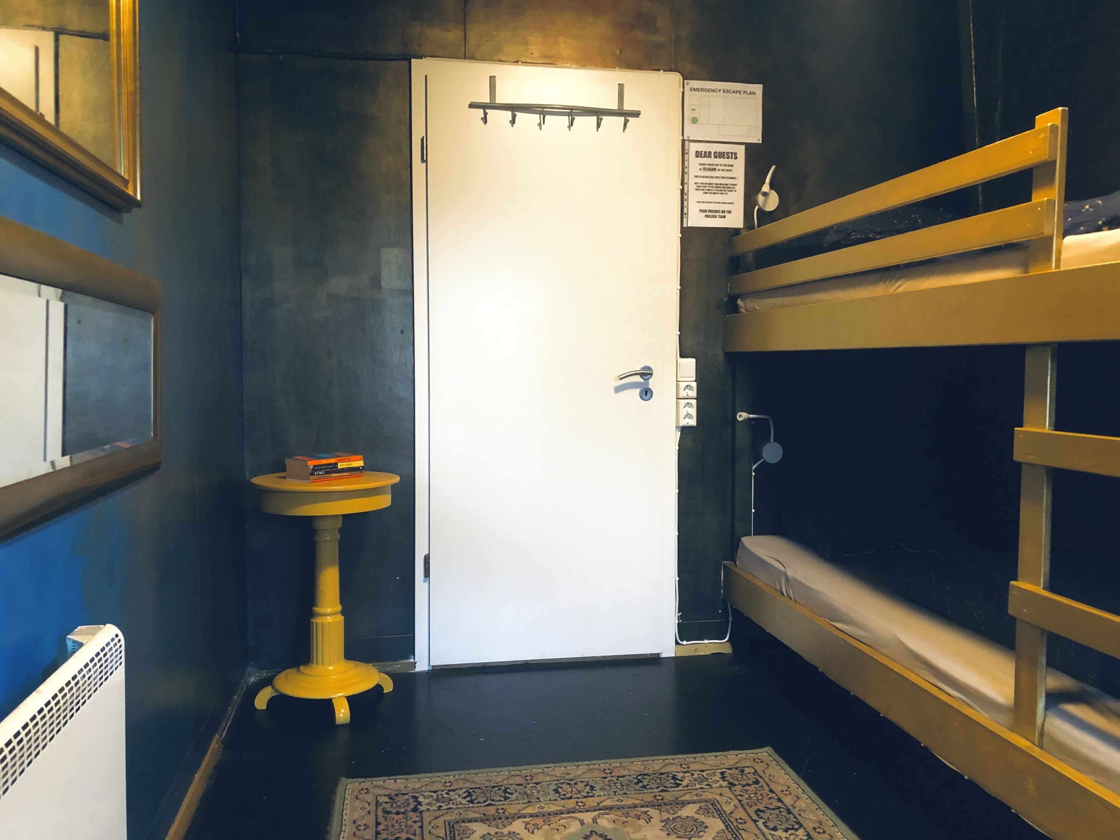 A compact room with a bunk bed, a small round table with books, a white door with a handle, and a wall-mounted emergency escape plan sign.