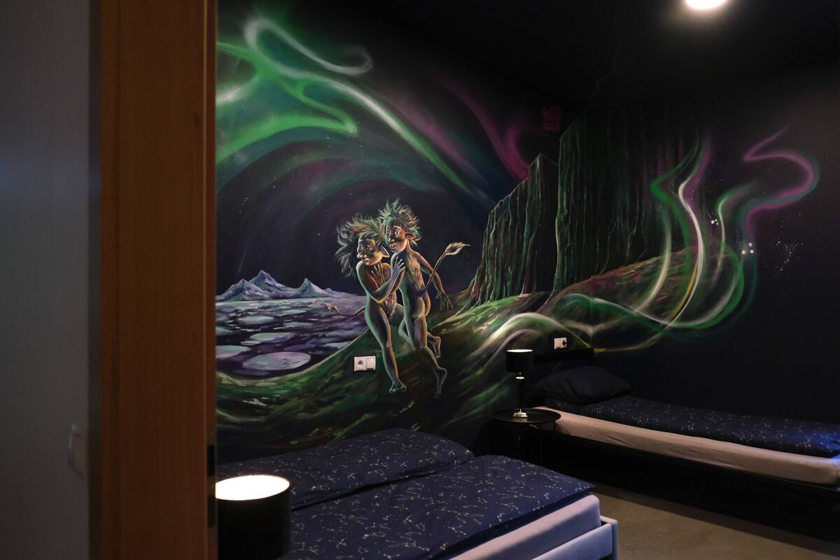 A room with a mural of a cosmic scene, a bed with a star-patterned cover, and a bedside table with a lamp.