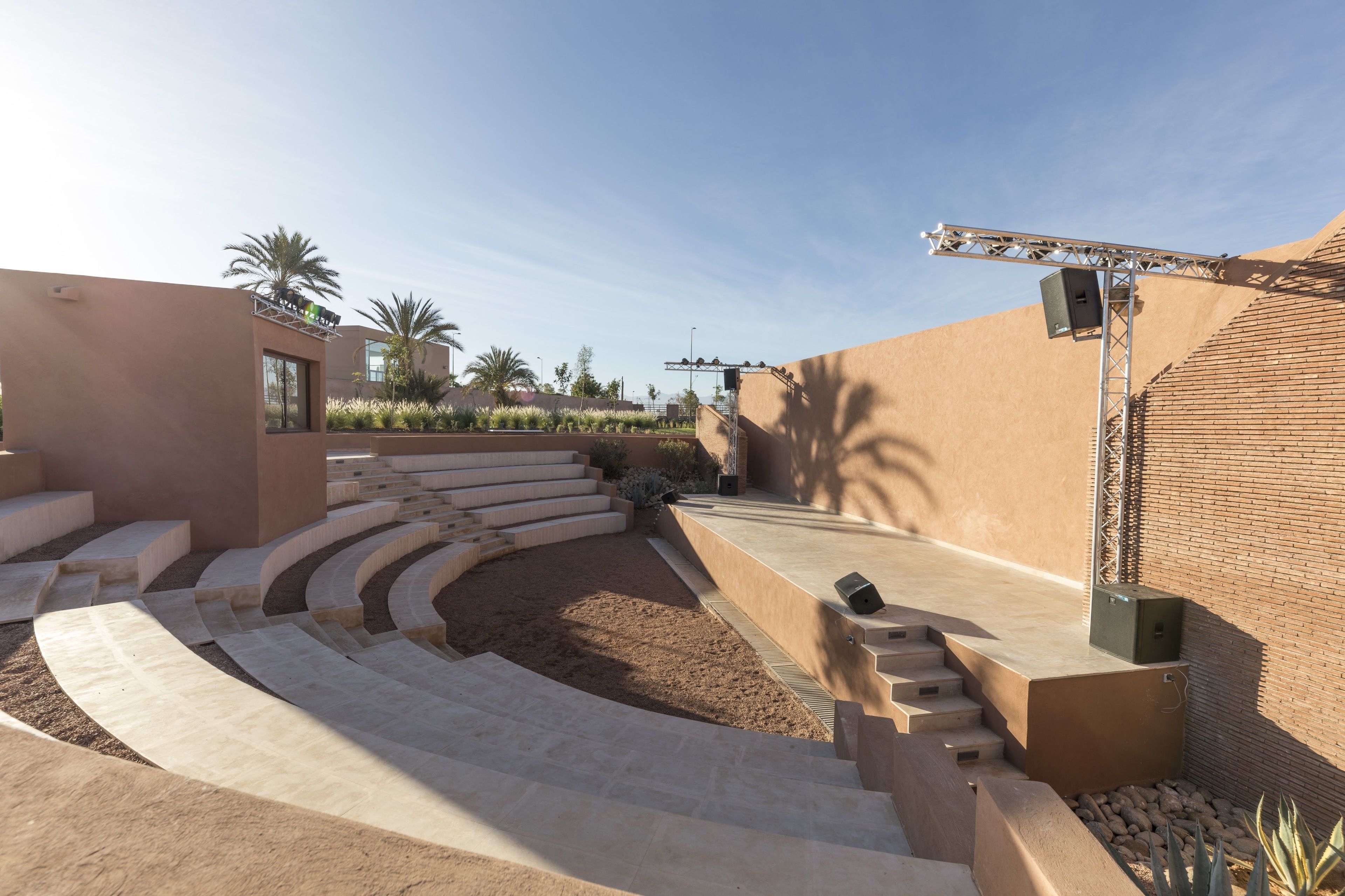 Un amfiteatre modern a l'aire lliure amb seients en terrasses i un escenari, envoltat de jardins ben cuidats i palmeres.