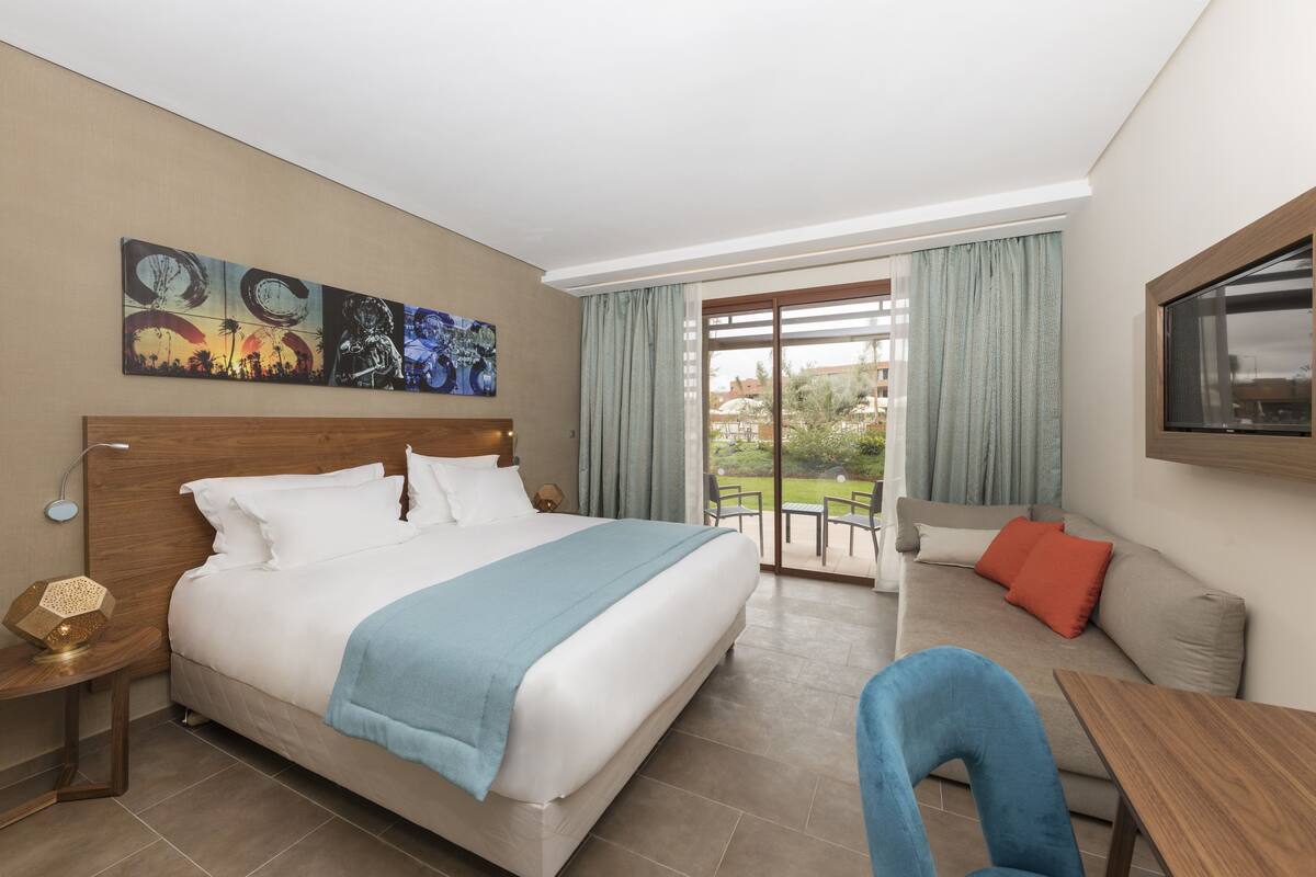 A hotel room with a large bed, a sofa, a dining table, and a balcony with a view of the outdoors.