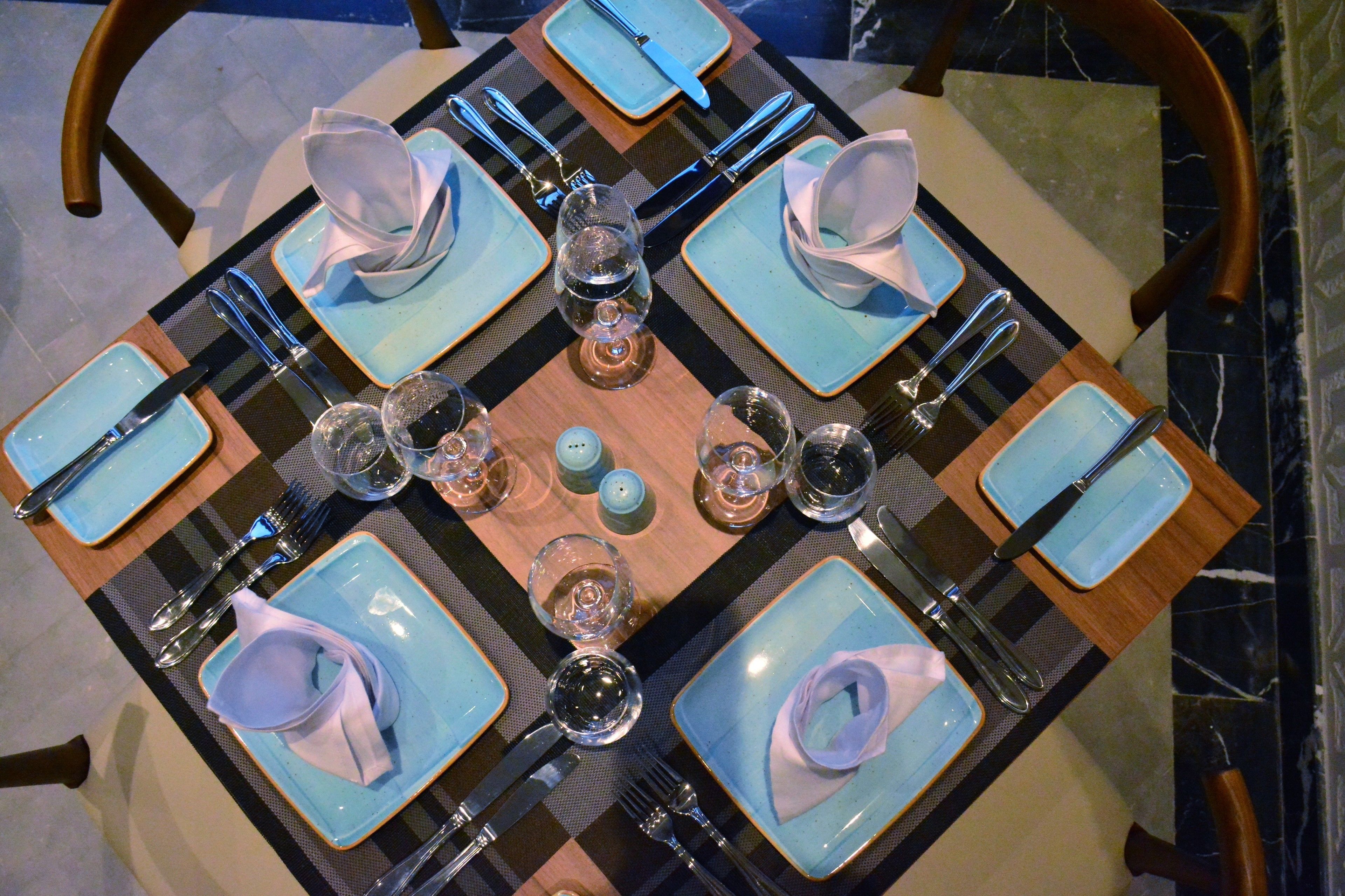 A square table with blue plates, silverware, and clear glasses, set on a checkered table runner.