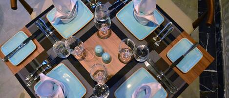 A square table with blue plates, silverware, and clear glasses, set on a checkered table runner.