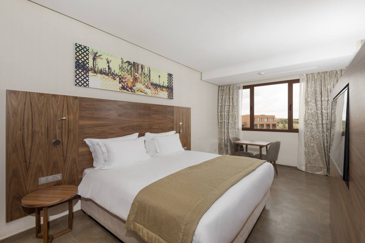A hotel room with a large bed, a wooden headboard, a small table, a chair, and a window with curtains.