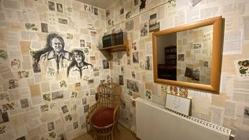 A room with a wall covered in various papers and photographs, a wooden chair with a red cushion, and a white radiator.