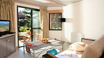 Un salon moderne avec un canapé, une table basse sur laquelle sont disposés des fruits, un téléviseur et un balcon offrant une vue sur les arbres et le paysage.