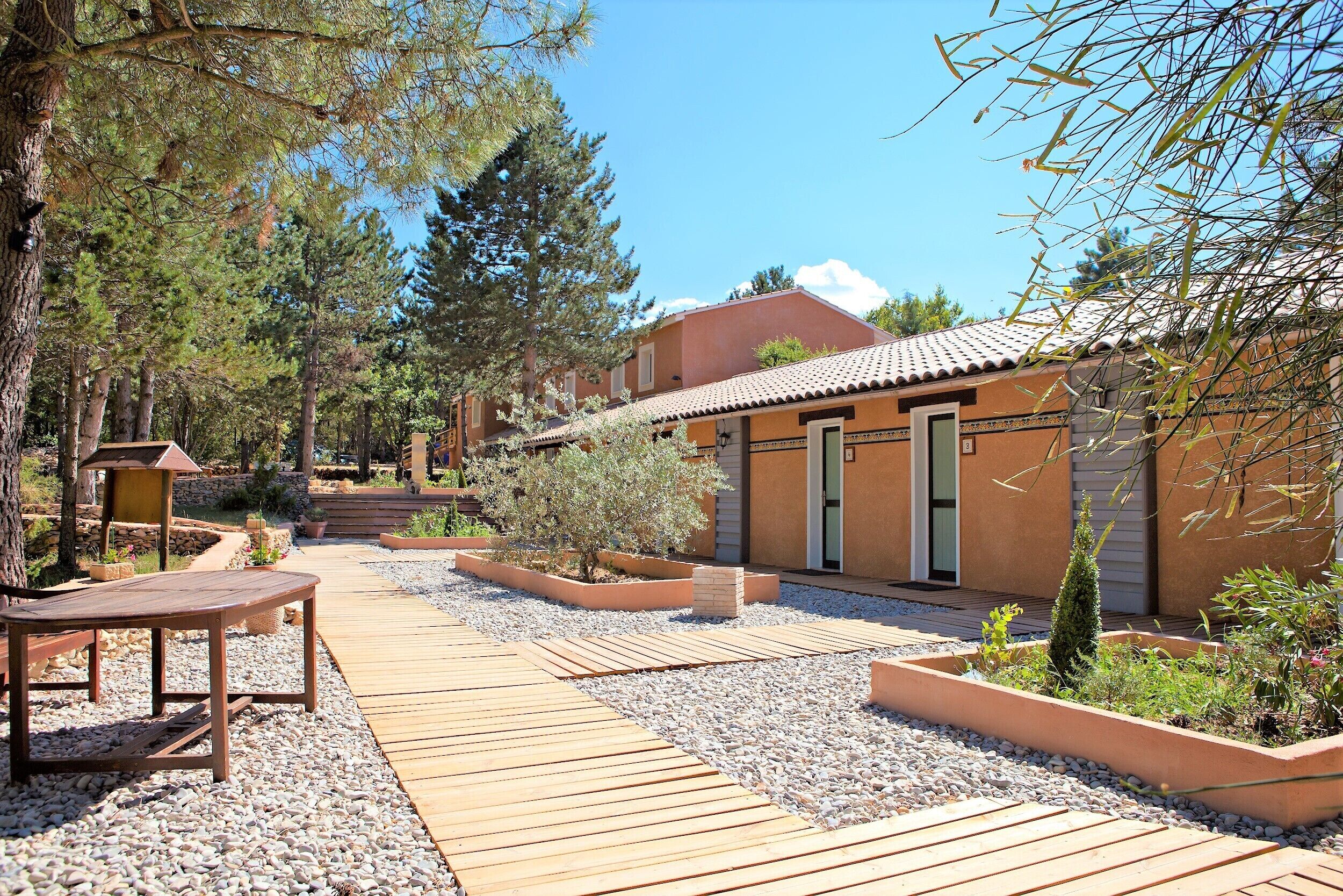 Un patio in legno con tavolo e panche, circondato da ghiaia e piante, che conduce a un edificio con tetto in tegole e una porta.
