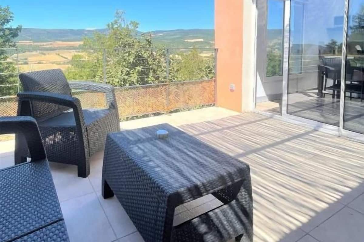A covered outdoor seating area with wicker furniture, a glass door leading to a patio, and a view of the countryside.