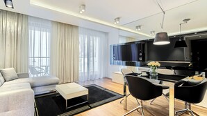 A modern living room with a sofa, dining table, and chairs. There is a TV mounted on the wall, and a kitchen area is visible in the background. - Imperial Apartments - Aquarius (Sopot)