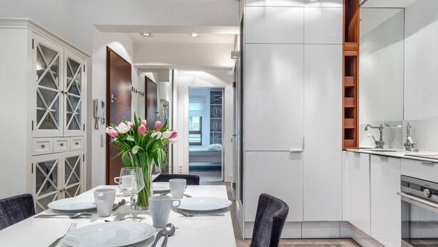 A modern kitchen with white cabinetry, a dining area with a table set for two, and a view into the bedroom. - Imperial Apartments - Aquarius (Sopot)