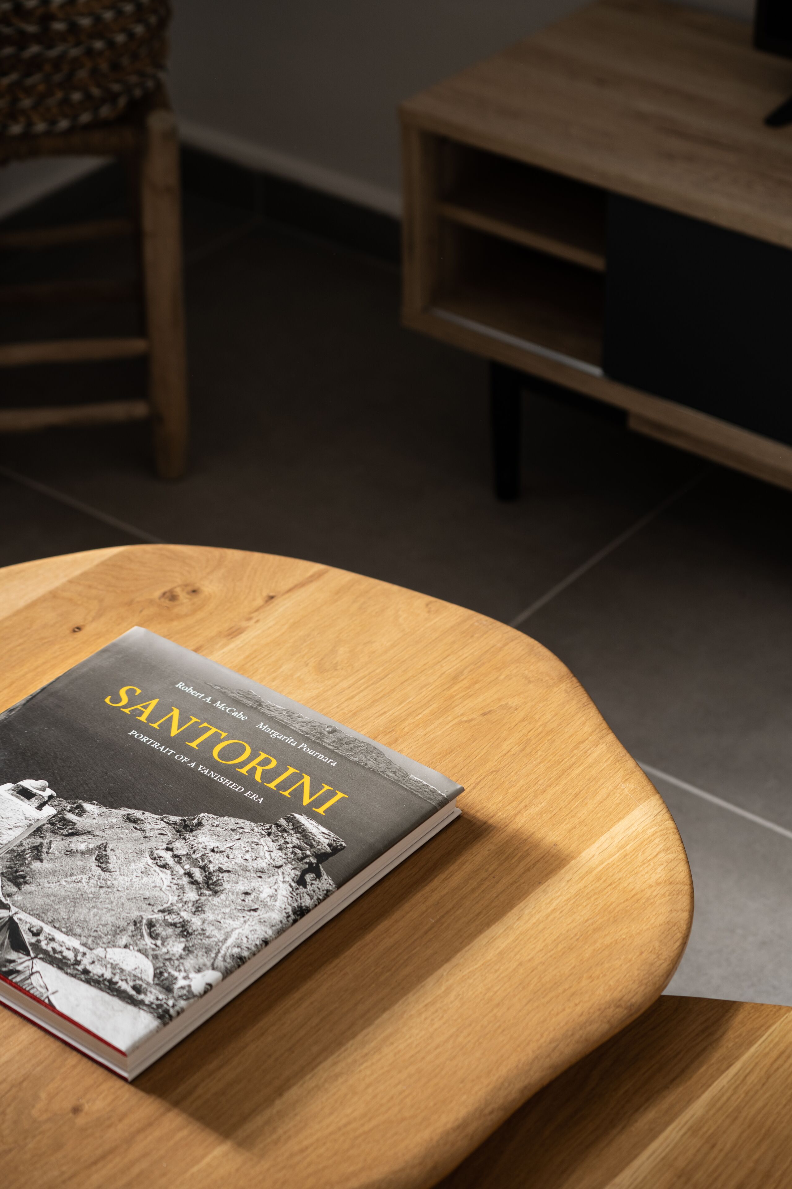 Een boek met de titel 'Santorini' op een ronde houten tafel in een kamer met een stoel en een plank.