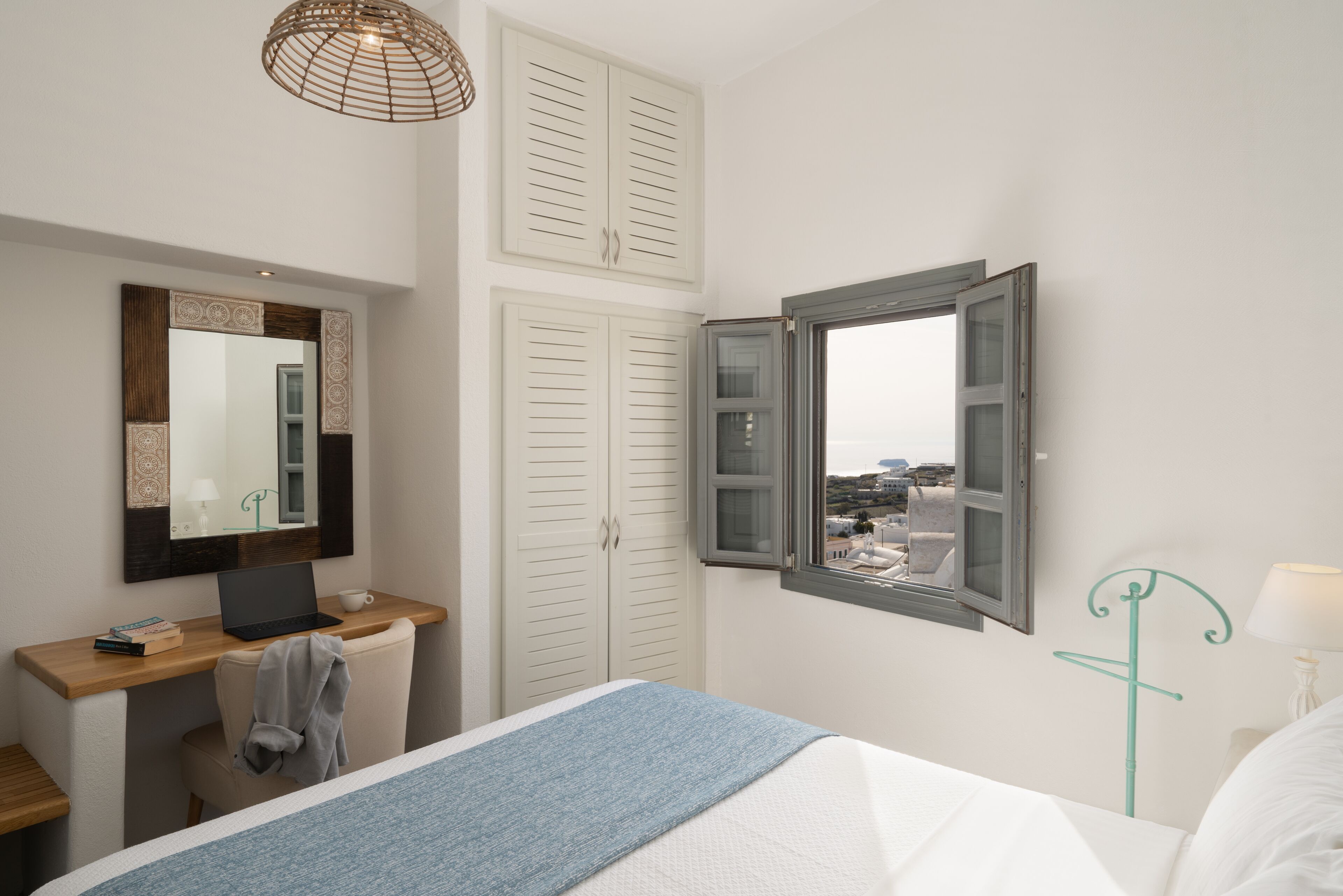 A bedroom with a bed, a desk with a laptop, a chair, a wardrobe, and a window with an open shutter.