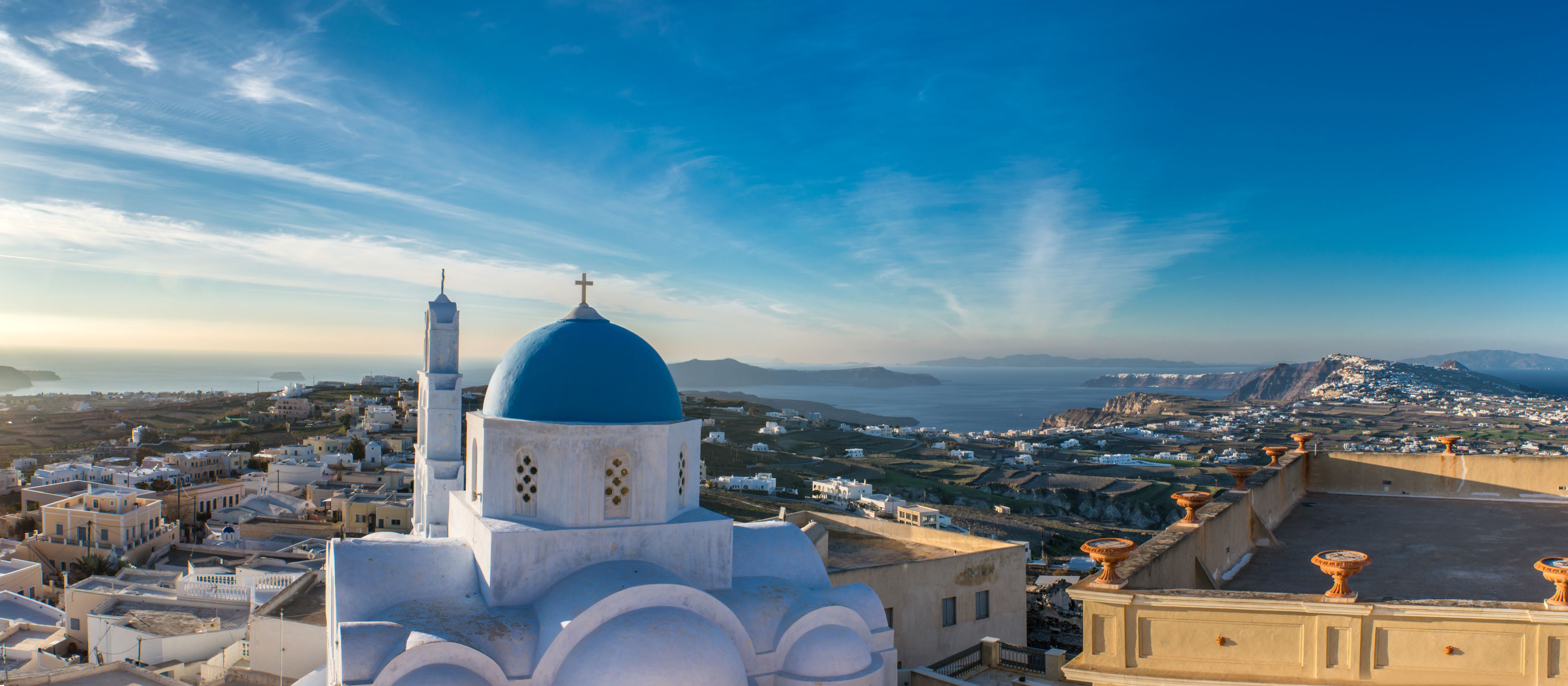 Un pueblo costero con edificios blancos y cúpulas azules, una iglesia destacada con una cruz, y vistas al mar y montañas distantes.
