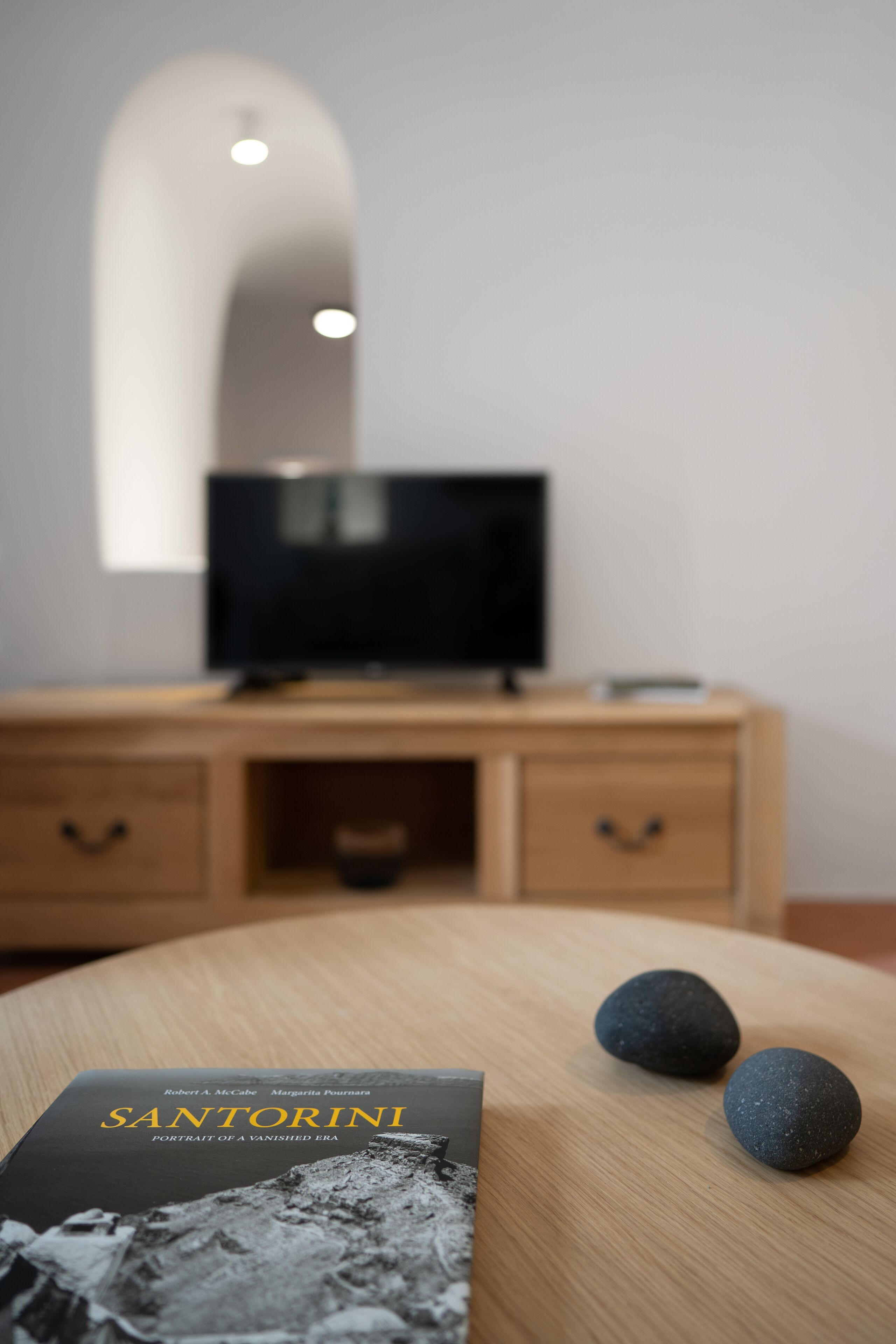 A room with a wooden TV stand, a flat-screen TV, and a book titled "Santorini" on a wooden table.