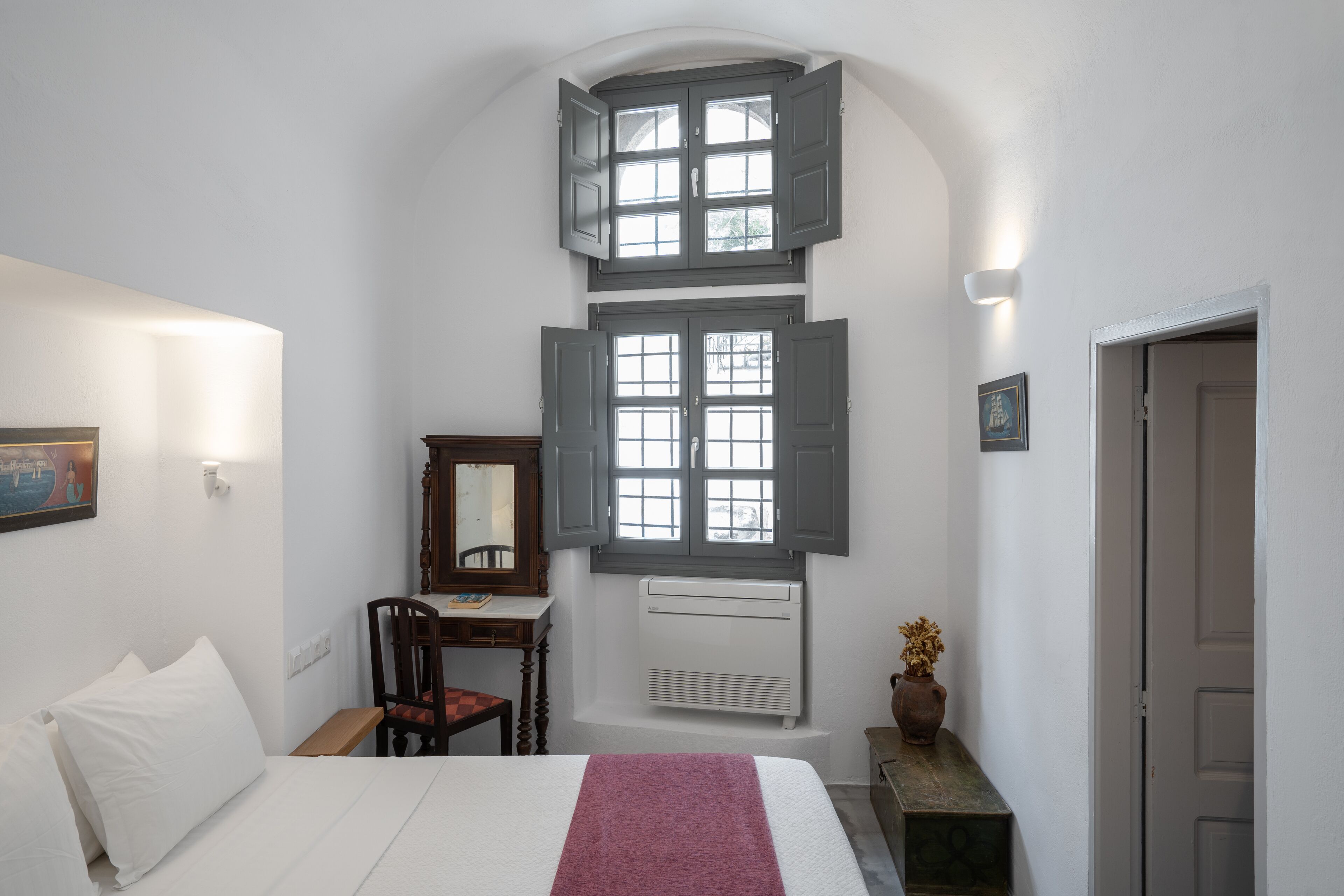 A bedroom with a bed, a desk, a chair, a window with shutters, a radiator, and a door.
