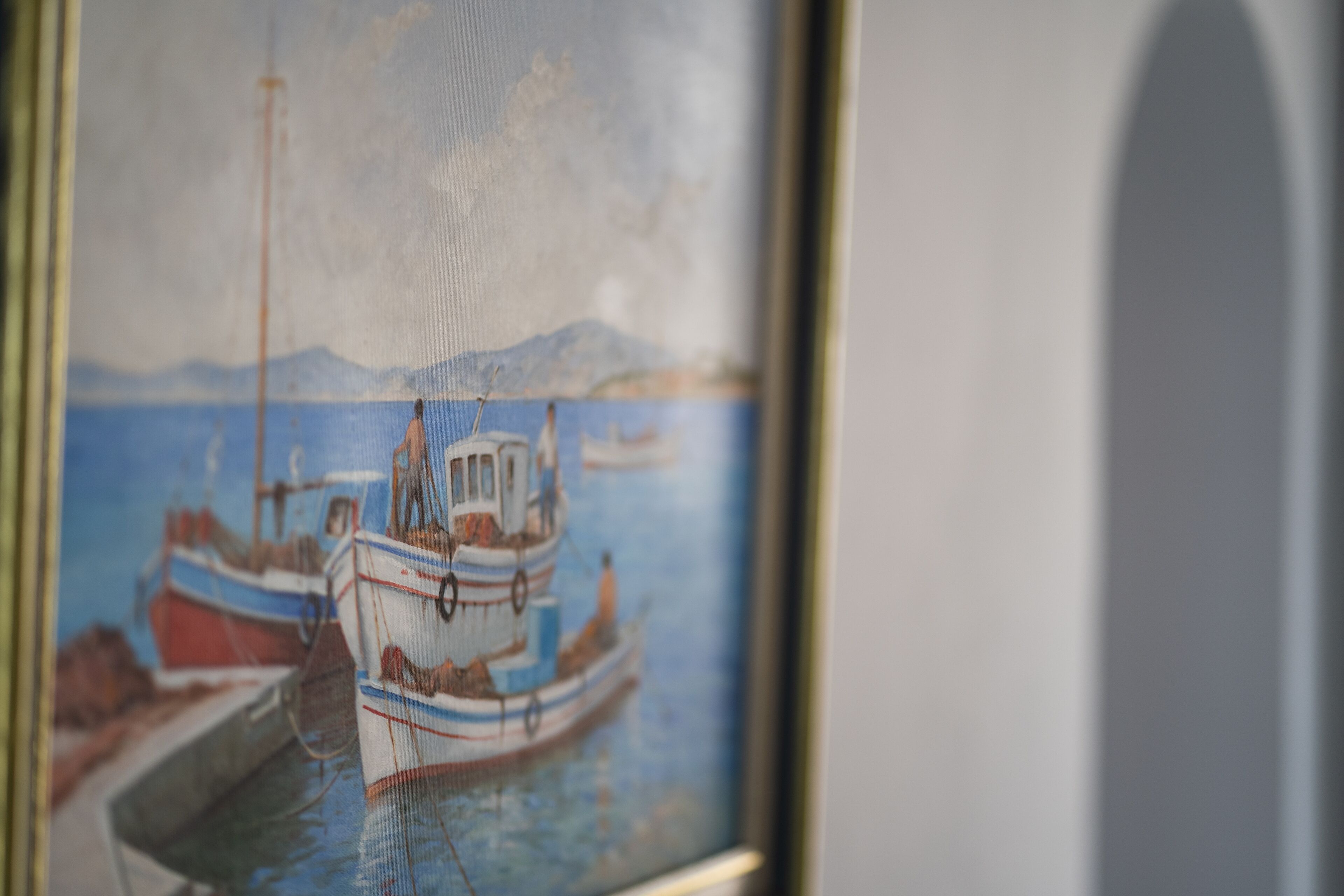 A framed painting of boats at a harbor with mountains in the background.