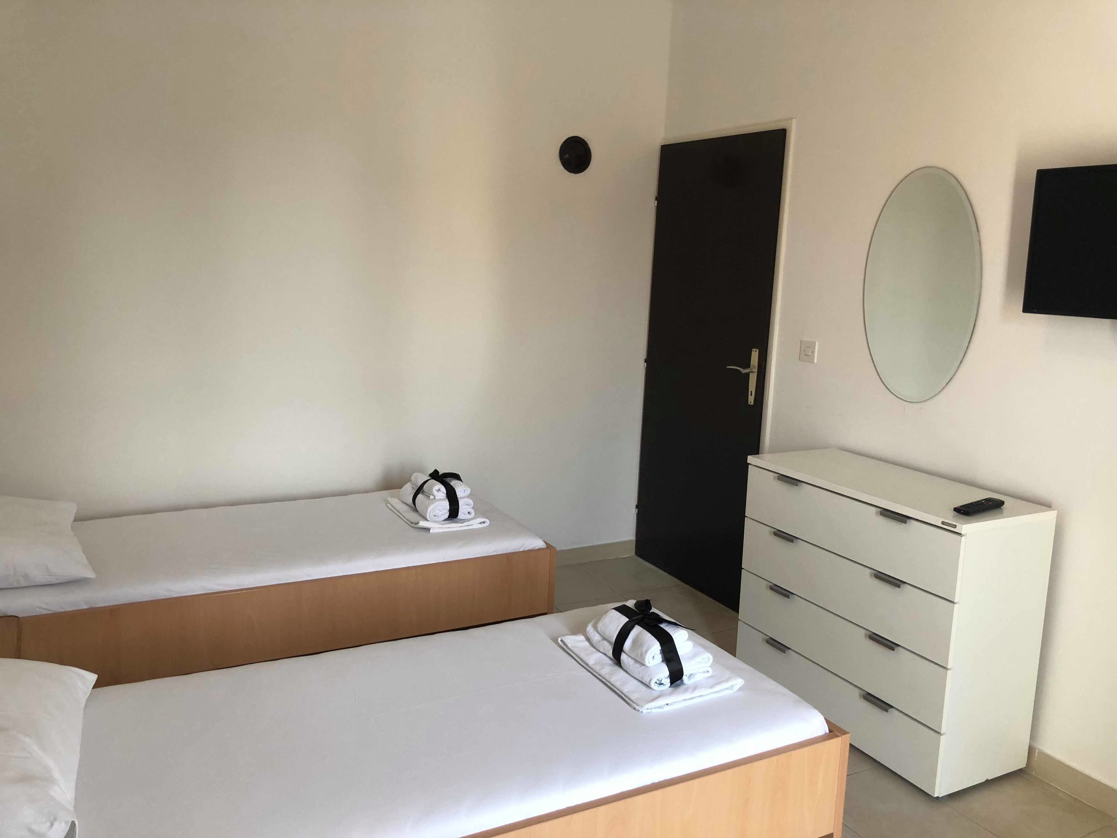 Hotellihuone, jossa on kaksi erillistä sänkyä, valkoinen lipasto, musta ovi, pyöreä peili ja seinälle asennettu televisio.