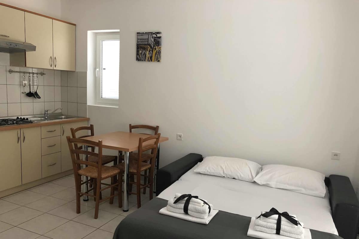 A compact hotel room with a kitchenette, a dining table with chairs, a bed with pillows, and a small window.