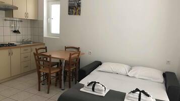 Quarto de hotel compacto com cozinha compacta, uma mesa de jantar com cadeiras, cama com travesseiros e uma pequena janela.
