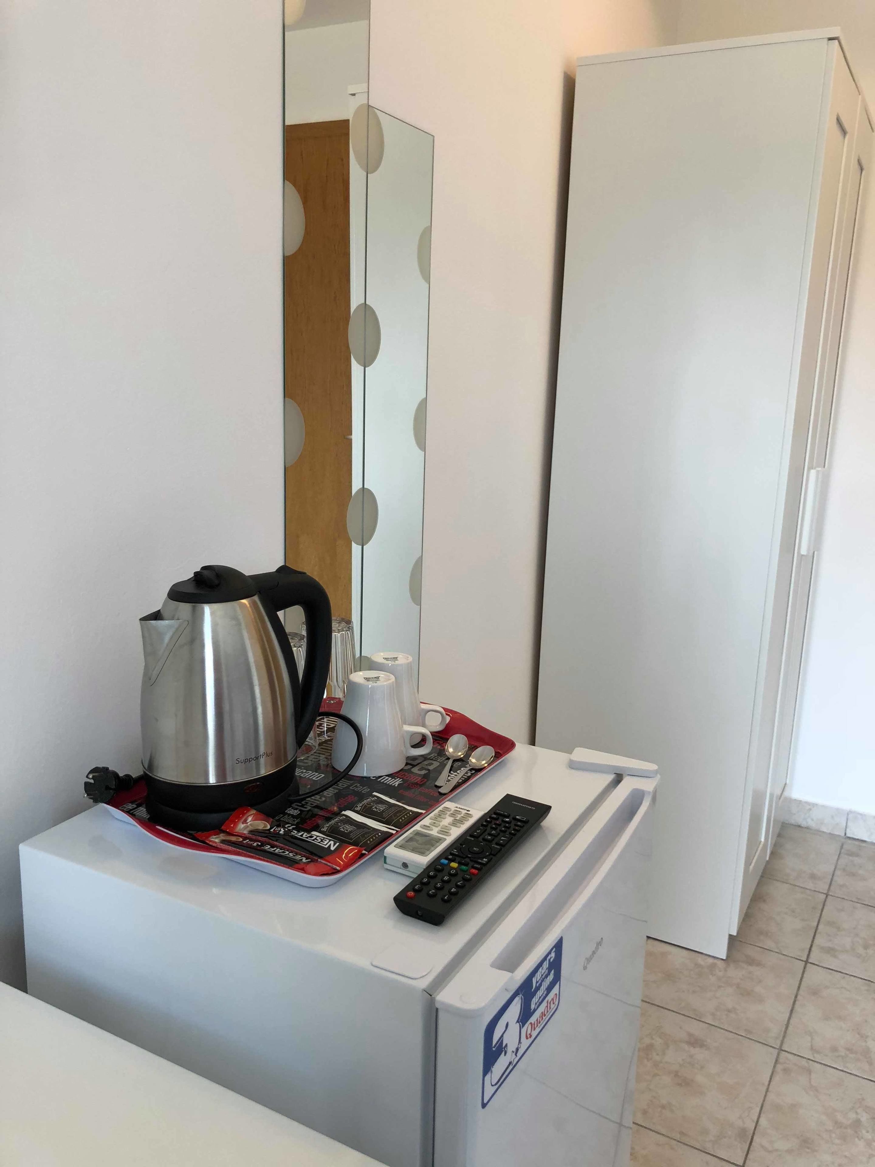 A compact kitchenette with a coffee maker, mugs, a tray, and a remote control on a small fridge.