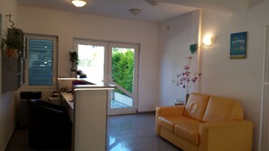 A modern living room with a yellow sofa, a kitchen area with a countertop, a dining table, and a large window with blinds. - Apartmani Ana&Ita (Novalja)