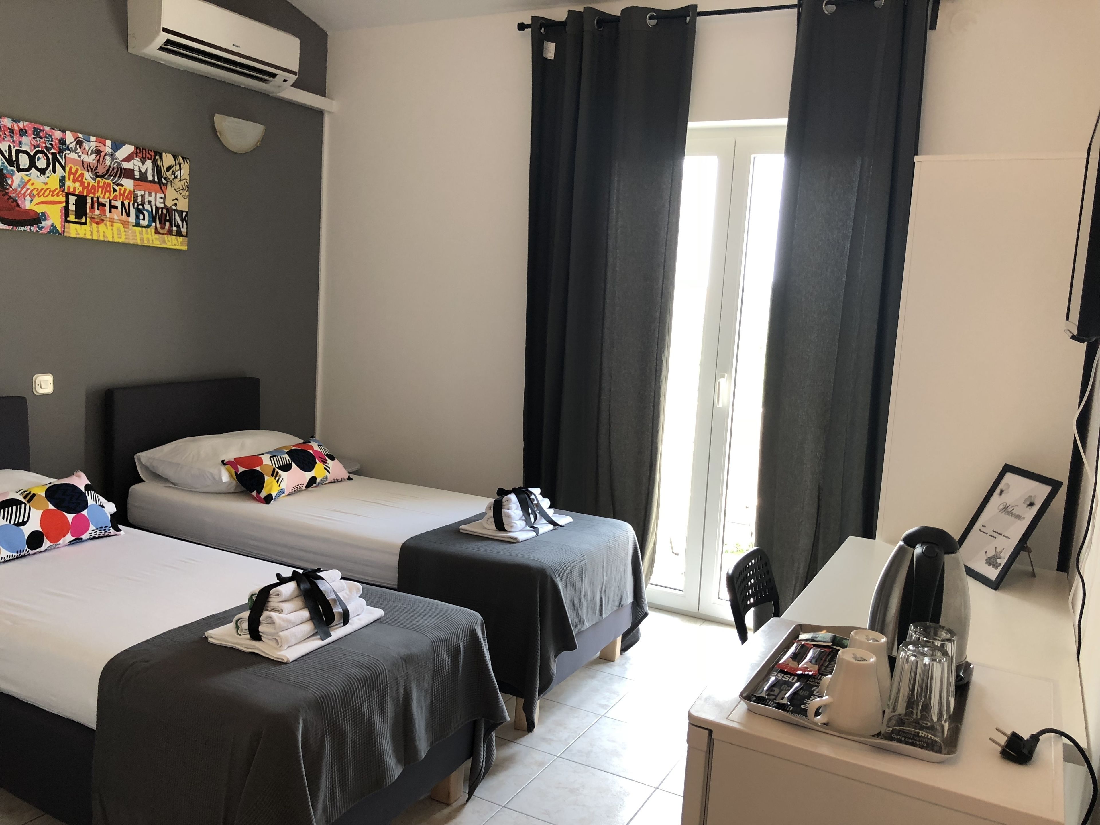 A hotel room with two beds, a desk with a kettle, and a window with curtains.