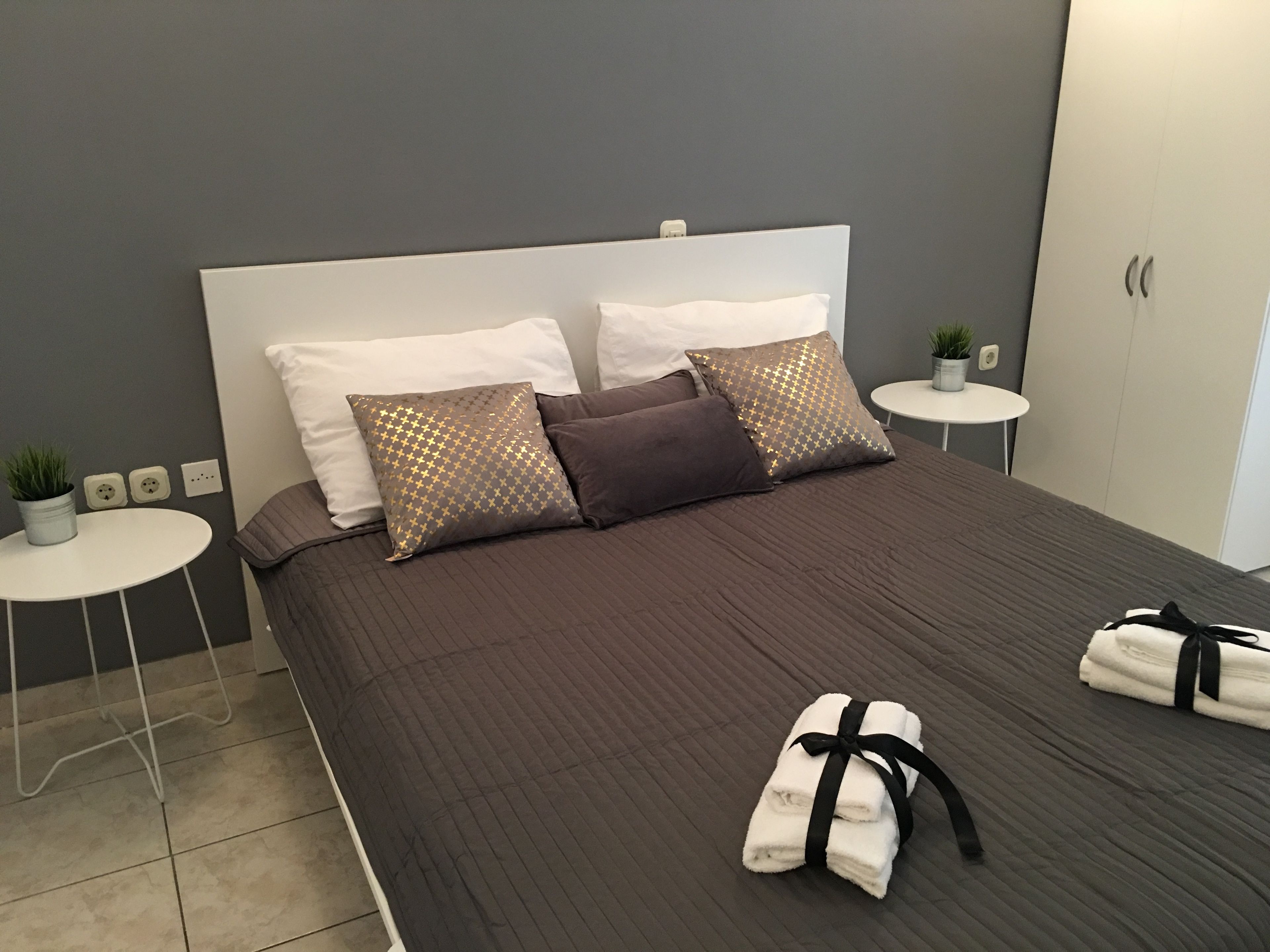 A neatly made bed with a brown quilt, two white pillows, and two patterned pillows. There are two small white side tables with plants, and a white wardrobe in the background.