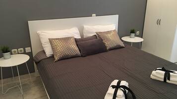 A neatly made bed with a brown quilt, two white pillows, and two patterned pillows. There are two small white side tables with plants, and a white wardrobe in the background.