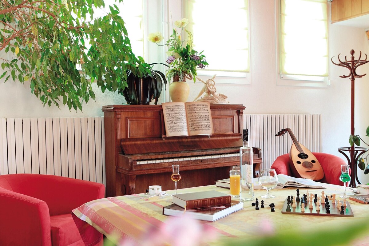 Um quarto acolhedor com um piano, uma guitarra, um tabuleiro de xadrez e uma mesa com livros e bebidas.