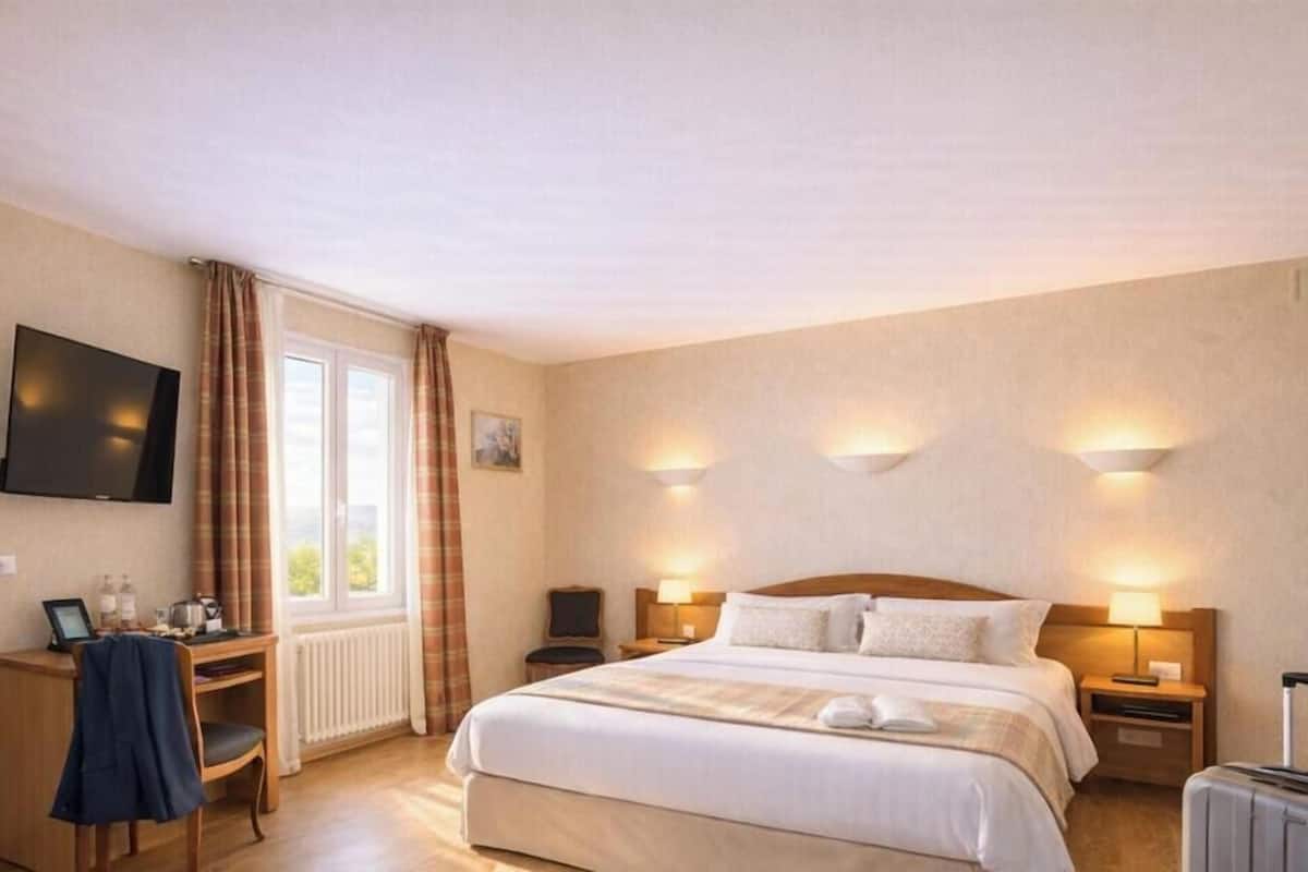Ein Hotelzimmer mit Bett, Nachttischlampen, Schreibtisch mit Stuhl, Wandfernseher und einem mit Vorhängen versehenen Fenster.