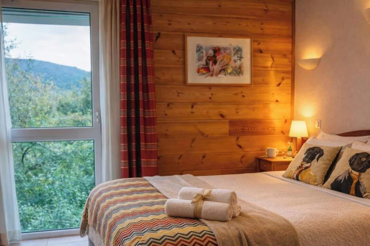 Ein Zimmer mit Holzwänden, einem Bett, einem mit Vorhängen versehenen Fenster und einer Wandlampe sowie einem gerahmten Bild.