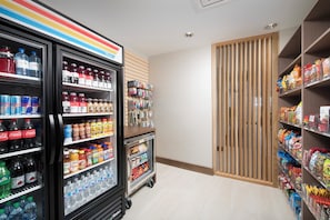 Interior de una tienda con dispensadores refrigerados para bebidas, estantes con snacks y una puerta de madera.