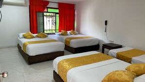 Quarto de hotel com quatro camas, cada uma com roupa de cama branca e detalhes dourados. Há uma janela com cortinas vermelhas, um aparelho de ar condicionado e um telefone na parede.