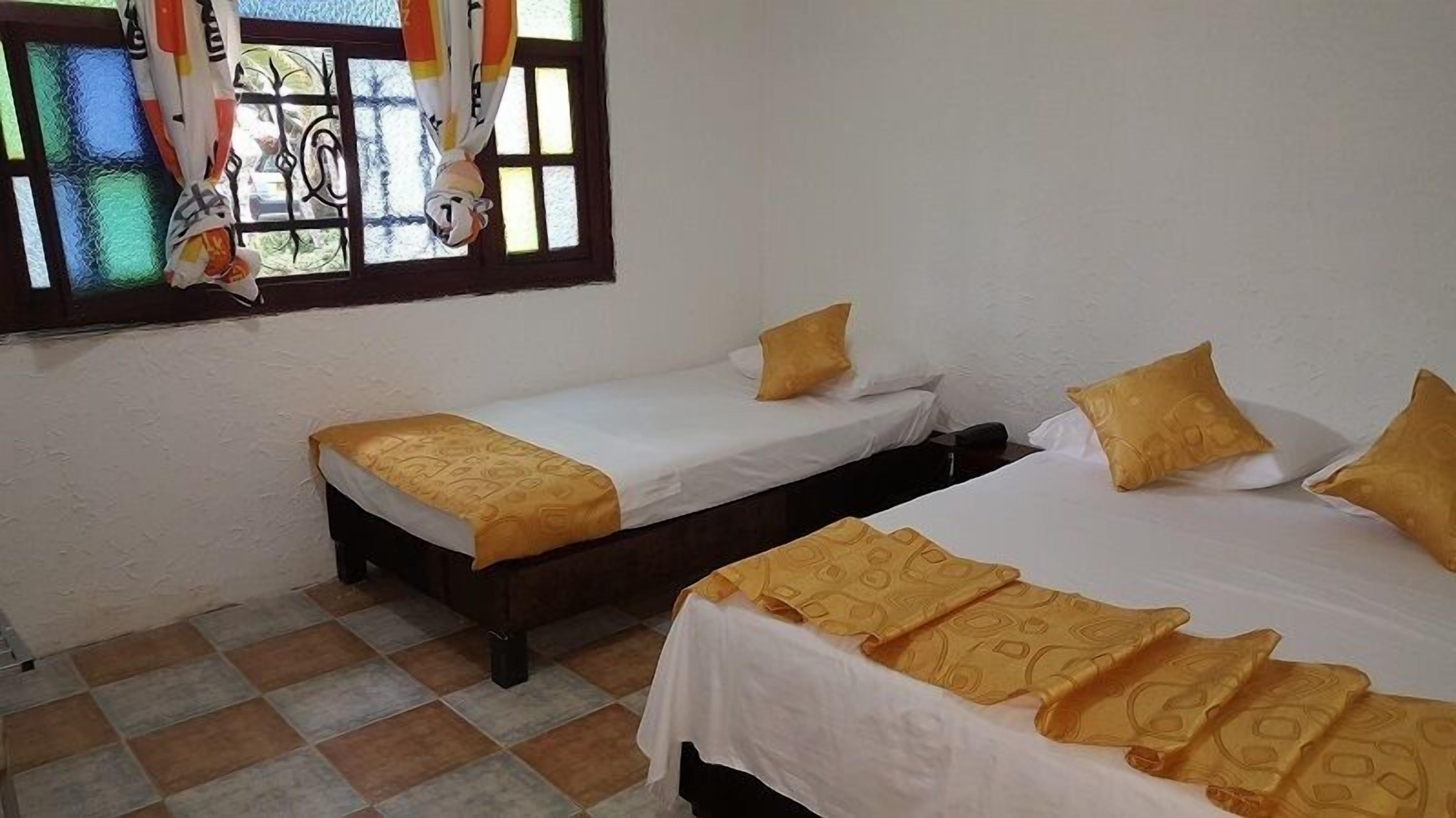 A room with two single beds, each with a white mattress and a patterned yellow bedspread. There is a window with a curtain, and the room has a tiled floor.