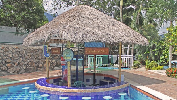 Uma área de piscina com um bar coberto por palha e um letreiro anunciando coquetéis e bebidas.