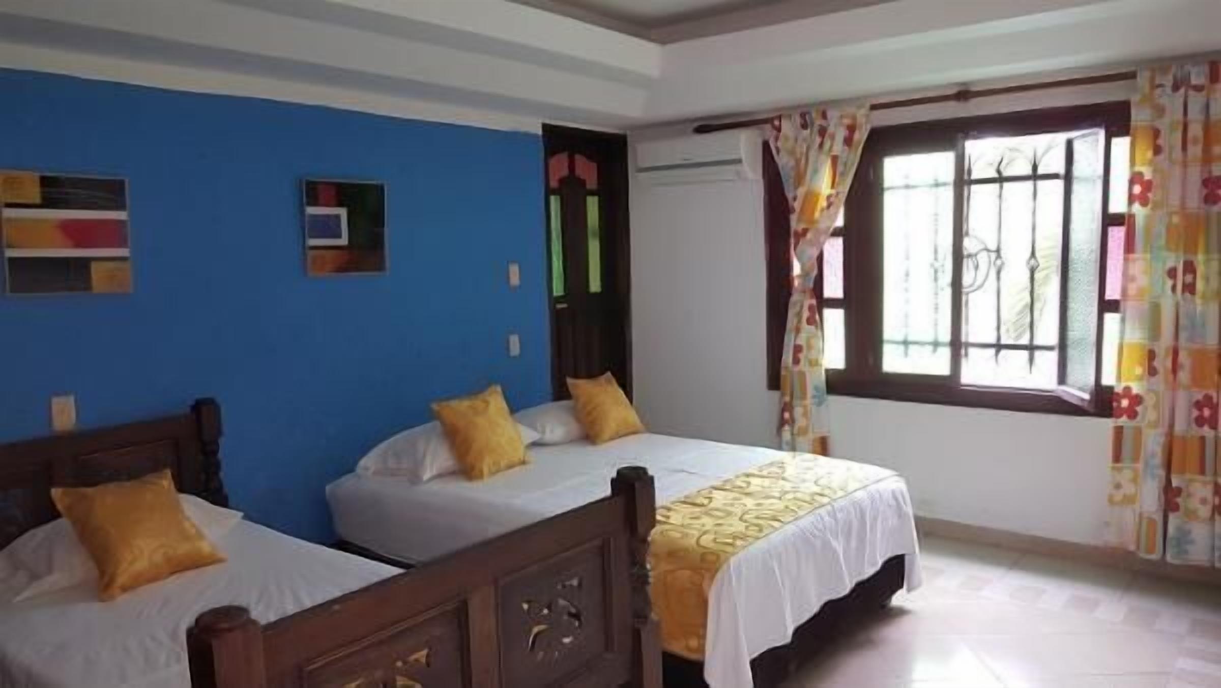 Una habitación con dos camas individuales, una pared azul, una ventana con cortinas y una puerta con marco verde.