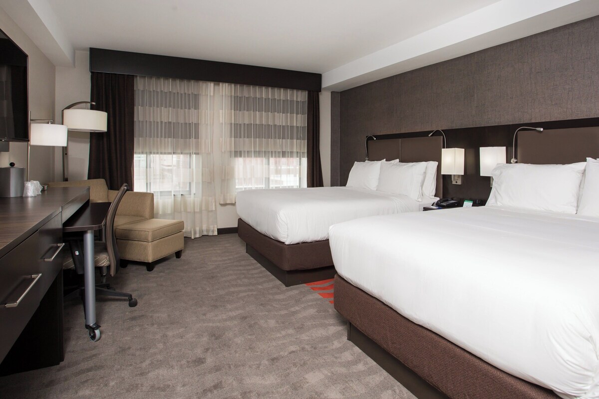 A hotel room with two beds, a desk, a chair, a large window with sheer curtains, and a flat-screen TV mounted on the wall.