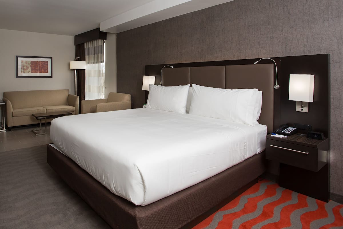 A hotel room with a large bed, a bedside table with a lamp, a nightstand, a desk, a chair, and a sofa.